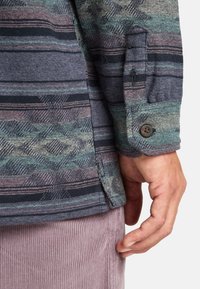 Chemise à boutons avec un motif géométrique multicolore dans des tons de vert, violet et noir; tissu texturé; manches retroussées avec détail boutonné.