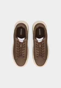 Pāris brūnu zamša GUESS kedu ar krēmkrāsas zolēm un brūnām auklām, skatīti no augšas uz balta fona.