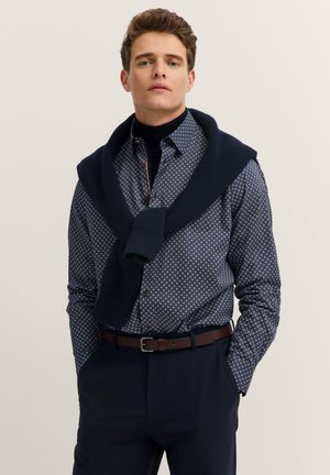 Camicia a pois blu navy e bianca da uomo con un maglione scuro annodato intorno alle spalle, abbinata a pantaloni scuri e una cintura marrone.