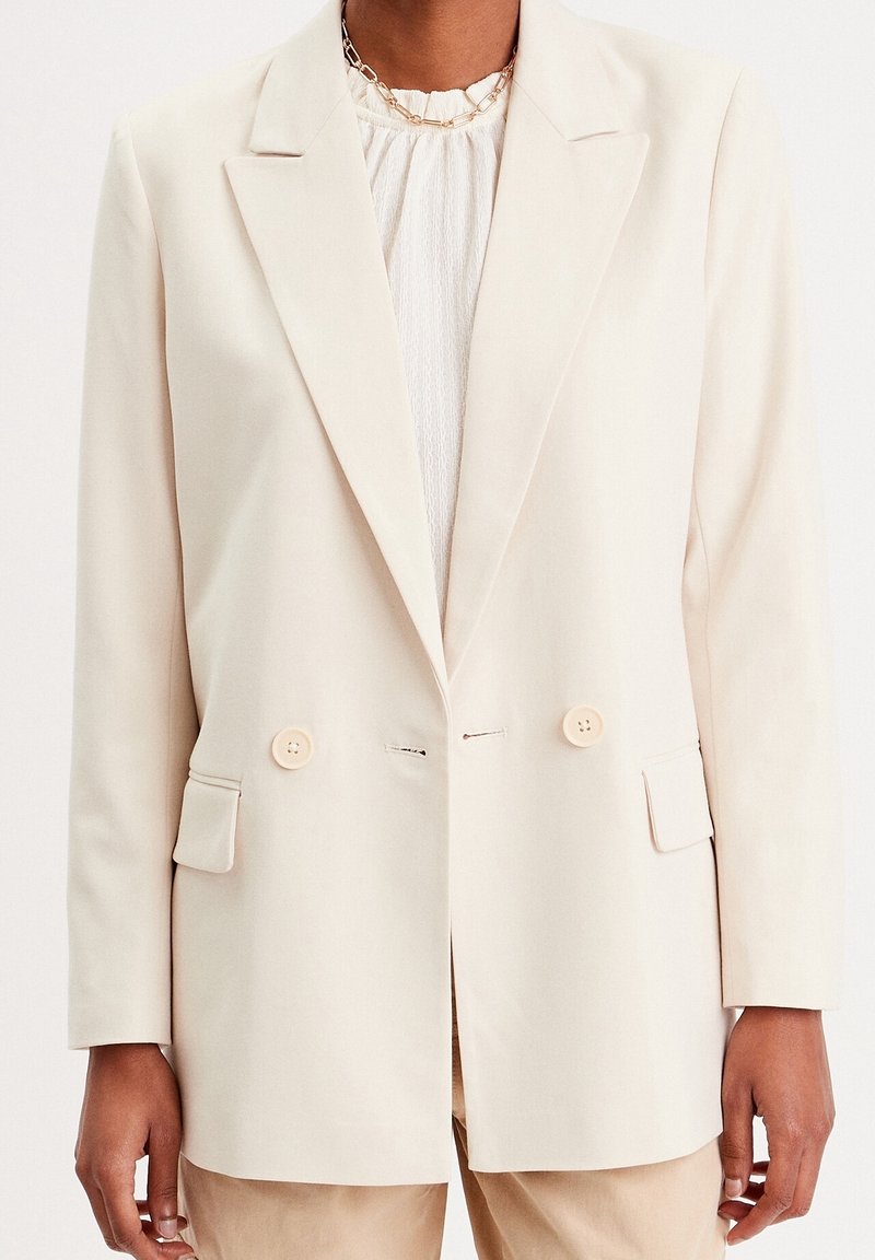 Lys beige dobbeltknappet blazer med klapkrave, to forlommer og blødt stof. Har runde knapper som pynt.