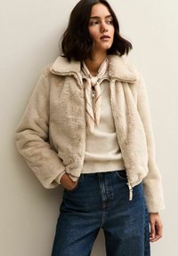 Chaqueta bomber de piel sintética en beige con cuello vuelto, cierre de cremallera y puños acanalados, combinada con un suéter claro y vaqueros oscuros.