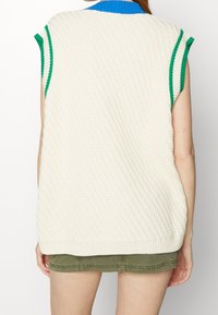 Stickad ärmlös tröja i krämfärg med diamantmönster. Har gröna och blå ribbade detaljer vid kragen och ärmhålen. Lätt oversized passform.