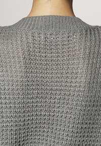 Back view of a person wearing a grey, loosely knitted sweater with a ribbed neckline, showing textured open weave pattern.
