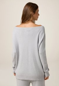 Pull en tricot gris clair avec un large décolleté à épaule dénudée, des poignets et un ourlet côtelés. Texture douce, coupe décontractée, légèrement plus long à l'arrière.