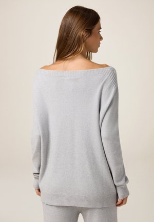 Pull en tricot gris clair avec un large décolleté à épaule dénudée, des poignets et un ourlet côtelés. Texture douce, coupe décontractée, légèrement plus long à l'arrière.