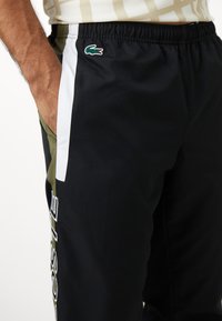 Svart treningbukser med grønne detaljer, elastisk midjebånd, sidelommer og hvite striper. Har en Lacoste-logo.