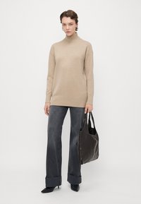 Femme portant un pull à col roulé beige, un jean large noir avec des ourlets, des bottes à talons noires, tenant un sac fourre-tout en cuir noir.