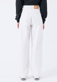 Jeans de mezclilla blancos, de pernera ancha y cintura alta, con bolsillos traseros y una etiqueta de cuero marrón en la cinturilla. Textura suave, diseño casual.