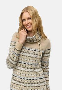 Pull beige à motifs avec un col en cowl, présentant des motifs géométriques en noir et blanc, et des accents de quincaillerie à cordon de tirage. Manches longues.