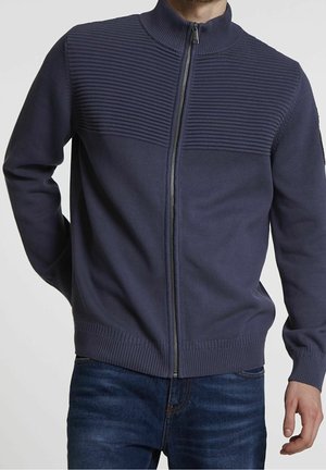 Mies, jolla on yllään laivastonsininen vetoketjullinen neulepaita, jossa on resoripintainen yläosa, sekä tummansiniset farkut, seisoo yksivärisen taustan edessä.