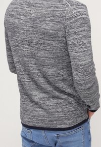 Pletený svetr s texturou v tmavě šedé barvě se světle šedými a bílými vzory. Má kulatý výstřih a žebrované manžety, s tmavě modrým lemem.