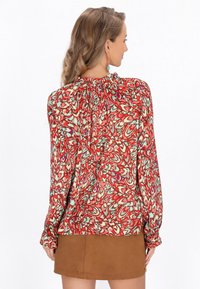 Løs rød bluse med et farverigt virvarmønster, lange ærmer, samlede udskæring og textureret stof; parret med en brun nederdel.