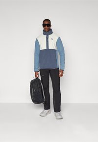 Pullover in pile blu e bianco con zip a un quarto, colletto alto e pattina con bottoni, abbinato a pantaloni neri e porta uno zaino nero.