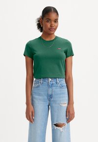 Groene katoenen T-shirt met korte mouwen en een ronde hals, voorzien van een klein rood logo op de borst. Gecombineerd met lichtblauwe versleten jeans.