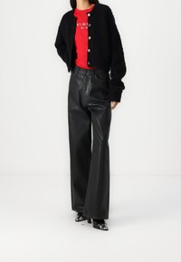 Cardigan noir duveteux avec boutons ronds, associé à un pantalon large en cuir noir brillant taille haute et des chaussures pointues noires avec des accents argentés.