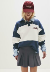 Junge Frau mit einer bordeauxfarbenen Illinois-Cap, einem blau-weißen Stockholm-Hoodie, einem gestreiften Hemd und einem dunklen Faltenrock vor einem weißen Hintergrund.