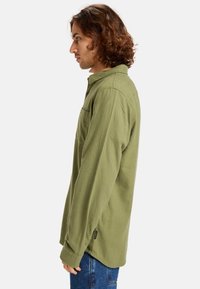 Camisa de manga larga verde oliva con cuello, bolsillo en el pecho y cierre de botones. Textura suave y ajustado ligeramente relajado.