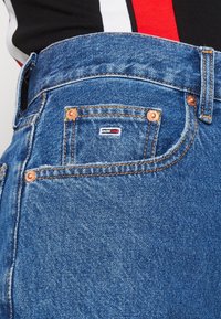 Modré džínové kalhoty s měděnými nýty, s malou logovou nášivkou v červené, bílé a modré barvě na zadní kapse. Klasický design s pěti kapsami.