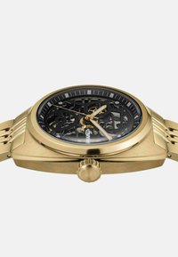 Gouden klok met een skeleton dial die ingewikkelde interne tandwielen onthult, zwarte bezel en een gestructureerde armband. Kronen bevindt zich op 3 uur.