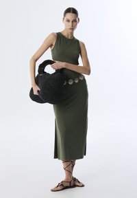 Olijfgroene mouwloze jurk met zijsplitten, een zwarte geweven tas met ronde handvatten, en zwarte sandalen met bandjes en decoratieve accenten.