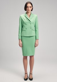 Blazer ajusté vert menthe avec double boutons et poches à rabat, associé à une jupe crayon ajustée, complété par des chaussures à talons noires.