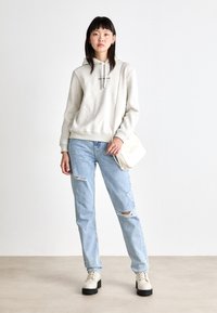 Lichtgrijze hoodie met een voorzak, gecombineerd met versleten lichtblauwe jeans. Het model draagt witte enkelboots en heeft een kleine crèmekleurige schoudertas.