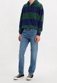 Sudadera a rayas verde y marino con tapeta frontal, combinada con jeans de color azul claro y zapatillas negras con suelas blancas.