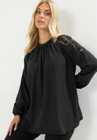 Schwarze Bluse mit puffierten langen Ärmeln, Spitzenverzierungen an den Schultern, rundem Ausschnitt und strukturiertem Stoff. Lässige Passform, abgeschlossen mit dekorativen Akzenten.