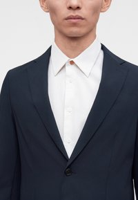 Homme portant une veste de costume bleu marine foncé sur une chemise blanche boutonnée jusqu'en haut avec un seul bouton marron visible.