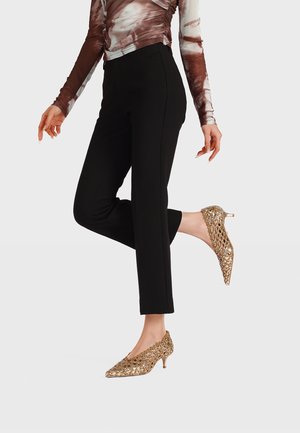 Donna che indossa pantaloni neri aderenti alla caviglia, maglia a maniche lunghe marrone e bianca e scarpe dorate con tacco basso a punta, in piedi su una gamba sola.