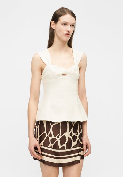 Γυναίκα με άσπρο αμάνικο μπλουζάκι με φαρδιές τιράντες και φούστα με καφέ και μπεζ ζωικό σχέδιο, στέκεται μπροστά σε απλό φόντο.