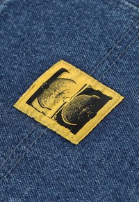 Tejido de mezclilla con una etiqueta dorada cosida que presenta un gráfico negro de dos caras superpuestas, sobre un fondo azul.