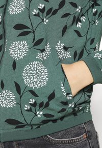 Grüne Blüten-zipper Jacke mit schwarz-weißen Löwenzahn-Mustern, strukturiertem Stoff und gerippten Bündchen. Hand-in-Tasche-Detail sichtbar.