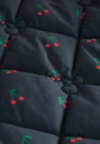 Gep quilteter schwarzer Stoff mit kleinen roten Kirschen und grünen Blättern, genäht in einem Rautenmuster, mit sichtbaren schwarzen Fadenakzenten.