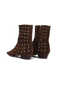 Bruine suede enkellaarsjes met lage houten hakken, bedekt met zilveren studs en zijritsluitingen, tegen een witte achtergrond.