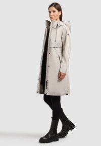 Ženska v svetlo bež dolg plašč s kapuco, črnih pajkicah in črnih masivnih škornjih, stoji ob enobarvnem ozadju.