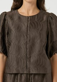 Brun textured bluse med bølge mønster, der har korte pufærmer og rund halsudskæring, lavet af et glat, shiny stof.