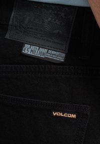 Zwarte denim jeans met een textuurlabel met reliëfgraphics, een label dat waterbesparing aangeeft, en subtiele gouden borduring van "VOLCOM."