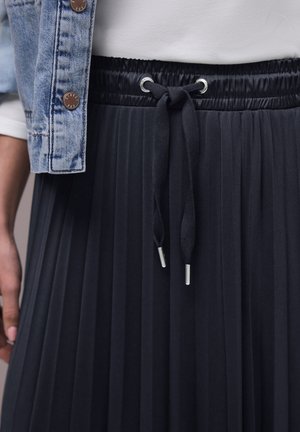 Plisseret sort nederdel med elastisk talje og bundet snor, kombineret med lys denimjakke og hvid skjorte.