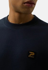 Granatowy sweter z ribbowanym dekoltem, z czarną kwadratową naszywką z złotym logo "PATRICK" na piersi.