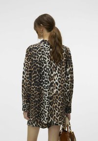 Překrývající se košile s leopardím potiskem v hnědé, černé a béžové barvě. Texturovaná tkanina s dlouhými rukávy a uvolněným střihem. Viditelný detail zapínání na knoflíky na zadní straně.