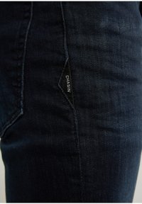 Tmavě modré džínové kalhoty s úzkým střihem, boční kapsou s černou štítkem s nápisem "CHASIN'" a hladkou, mírně vybledlou texturou.