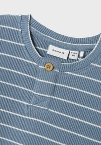 Maglia blu a coste con strisce bianche, caratterizzata da un collo rotondo, una patta frontale con un bottone in legno e un'etichetta che indica la taglia 9-12 mesi.