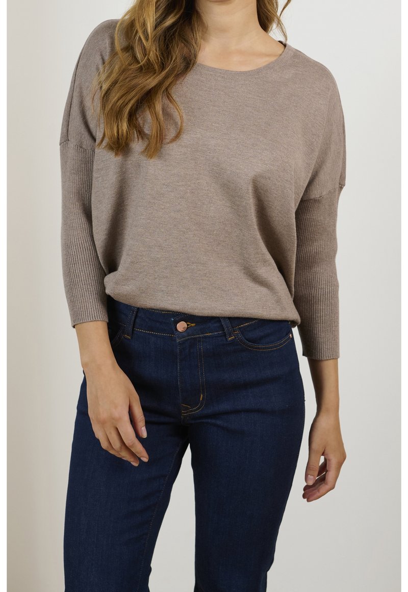 Femme portant un pull en maille taupe à manches longues avec des manches côtelées et un jean bleu foncé, debout devant un fond uni.