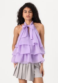 Lavendelfarget halterneck topp med lagdelte volanger, med en sløyfe rundt halsen. Stoffet er lett og noe gjennomsiktig, og kommer sammen med en grå plissert skjørt.