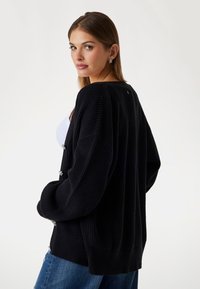 Black knitted cardigan with a loose fit, ribbed texture, and large buttons. Features dropped shoulders and a slight oversized silhouette.