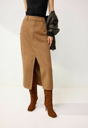 Femme portant une jupe midi taille haute beige avec une fente devant, des bottes en suède marron montant aux genoux, un haut noir et tenant une veste en cuir noire.