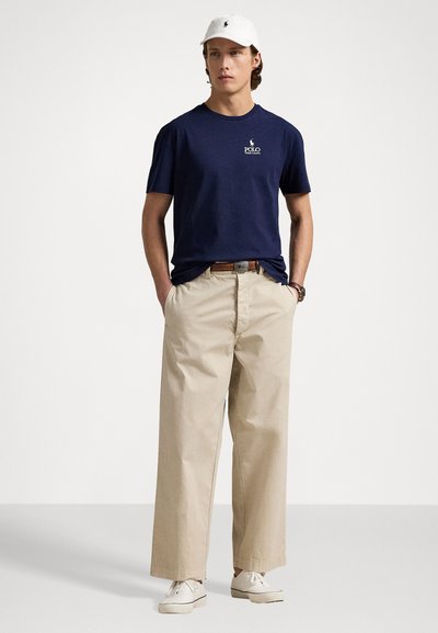 Navyblauwe katoenen t-shirt met een wit logo, beige wijdvallende chinos en witte sneakers. Heeft ook een witte cap. Eenvoudig ontwerp.