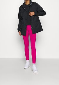 Svart lätt jacka med flera fickor över en åtsittande svart topp och ljusrosa leggings. Vita sportskor fulländar outfiten.
