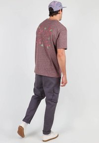 T-shirt bordeaux à manches courtes avec un motif géométrique vert et rose au dos, associé à un pantalon cargo gris et des baskets blanches.